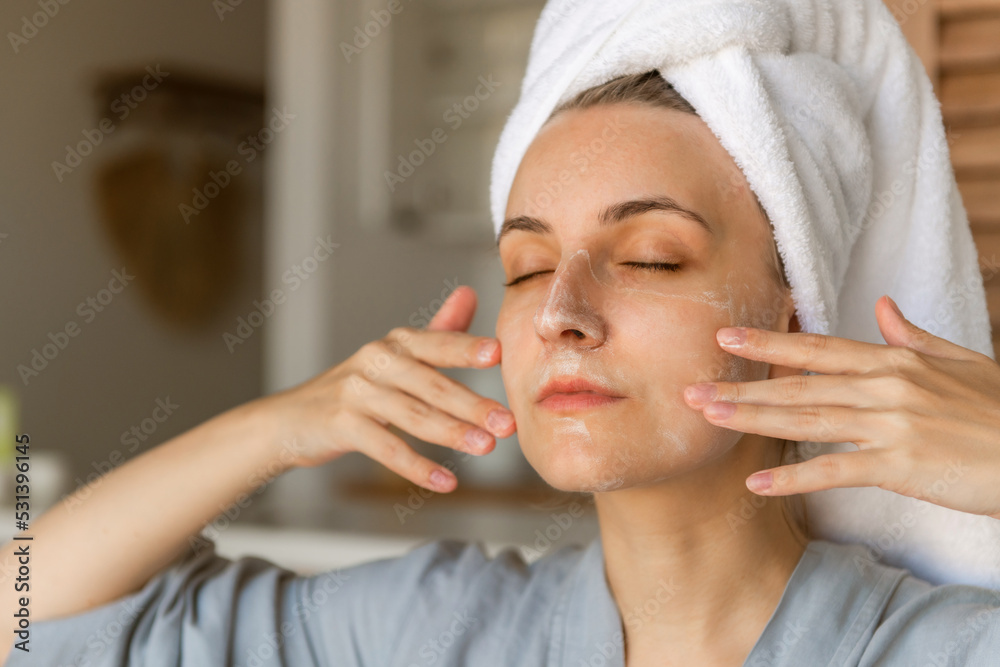 a young woman in a blue bathrobe and with a towel on her head sits on the bed after a shower. Woman taking care of her face skin with moisturizer. Skin care concept