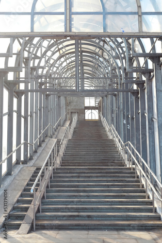 A staircase leading to a large covered bridge across the street. Perspective, arches, concrete, metal and glass.