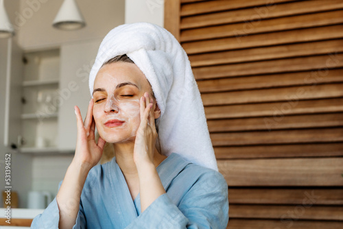 a young woman in a blue bathrobe and with a towel on her head sits on the bed after a shower. Woman taking care of her face skin with moisturizer. Skin care concept