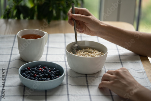 Morning meal. Female eating healthy breakfast with oatmeal porridge with summer berries - cowberry, blueberry, slice of butter, herbal tea. Clean eating, dieting, weight loss. Shallow depth of field