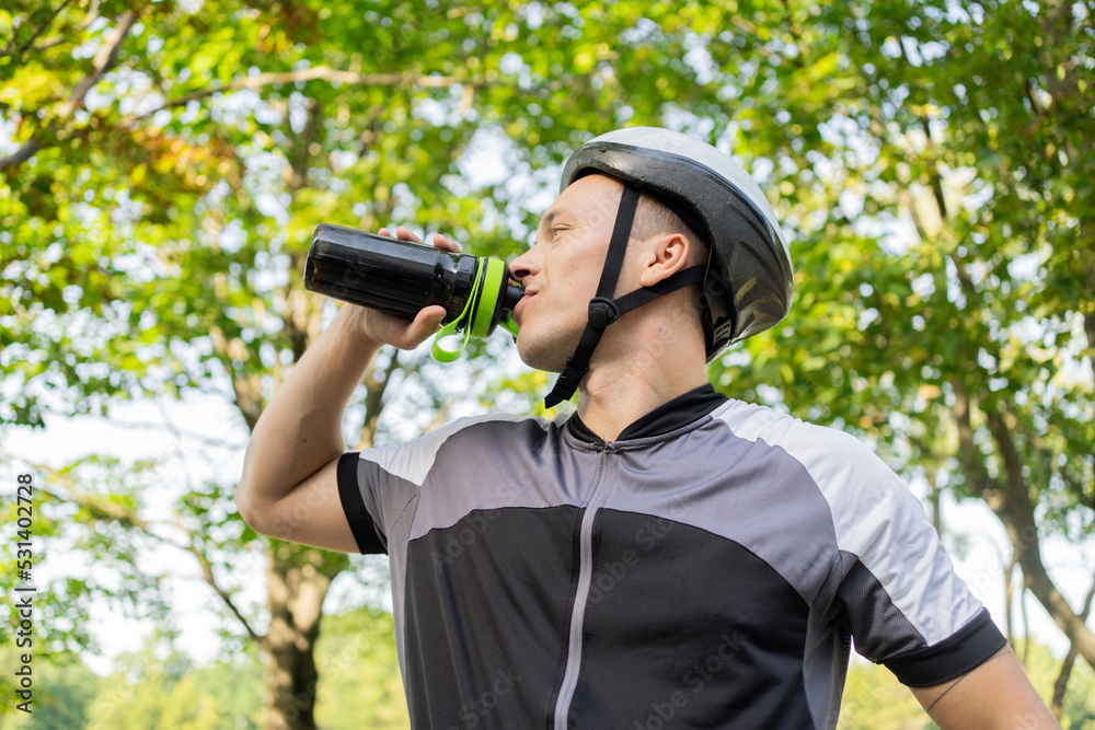 A male cyclist in a helmet drinks water from a bottle. A male cyclist in a helmet drinks water from a bottle.