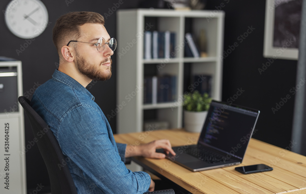 Young man coder thoughtful, sitting near desk with laptop in dark office, working on a innovate software Young man coder thoughtful, sitting near desk with laptop in dark office, working on a innovate software