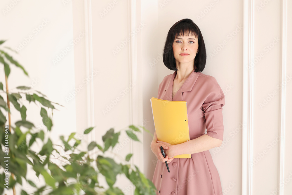 Attractive young woman in stylish soft pink dress is holding yellow folder of paperwork with mock up area looking in camera and standing in office with houseplants. Education, business, lifestyle Attractive young woman in stylish soft pink dress is holding yellow folder of paperwork with mock up area looking in camera and standing in office with houseplants. Education, business, lifestyle