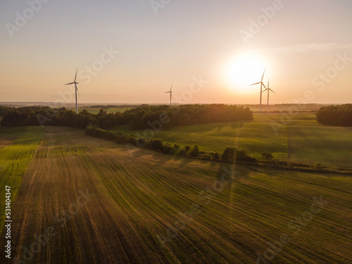 Green energy concept. Aerial cinematic drone panoramic shot of windmills and electricity wind turbines farm during summer sunset. Panoramic European nature view. Image contain noise and blur