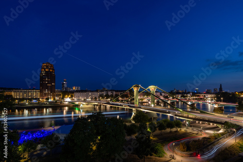 Bustling, vibrant nightlife in Frankfurt, Germany, with Flösserbrücke bridge in the foreground, and multiple trailing lights of vehicles and boats
