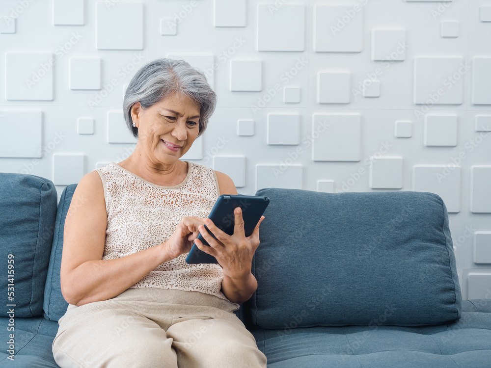Happy Asian senior woman sit on couch, holding and looking at digital tablet screen in hand in living room. Elderly female using smart device, computer at home. Older people use technology with easy. Happy Asian senior woman sit on couch, holding and looking at digital tablet screen in hand in living room. Elderly female using smart device, computer at home. Older people use technology with easy.