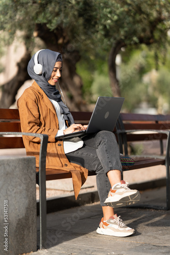 Muslim woman using a laptop and headphones outdoors