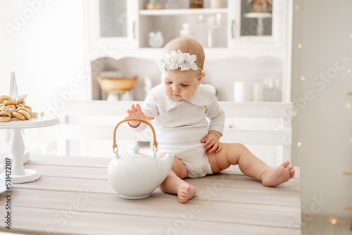 a happy baby in a white cotton bodysuit is sitting on the kitchen table with a hot kettle in a bright kitchen, a small smiling baby boy or girl is eating
