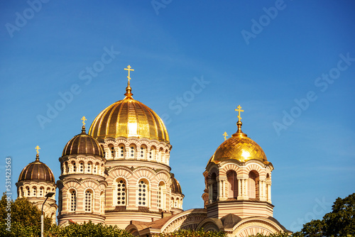 A church with a yellow wall and a pointed golden tip in the middle of the city center with multiple windows and trees next to it