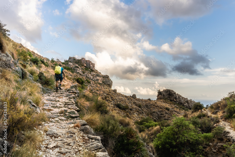 Hiker ascending the mountain along a cobblestone path, with a cloudy blue sky in the background