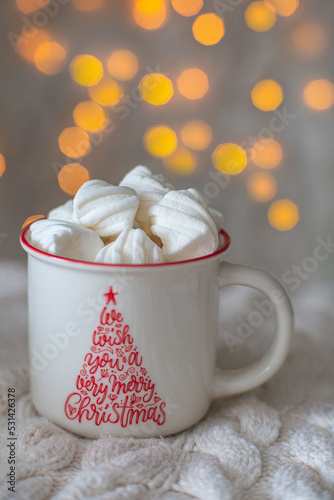 Hot chocolate with marshmallows in white mug, copy space. Hot cocoa drink for Christmas and winter holidays with warm scarf, lights on white table. We wish you a merry christmas