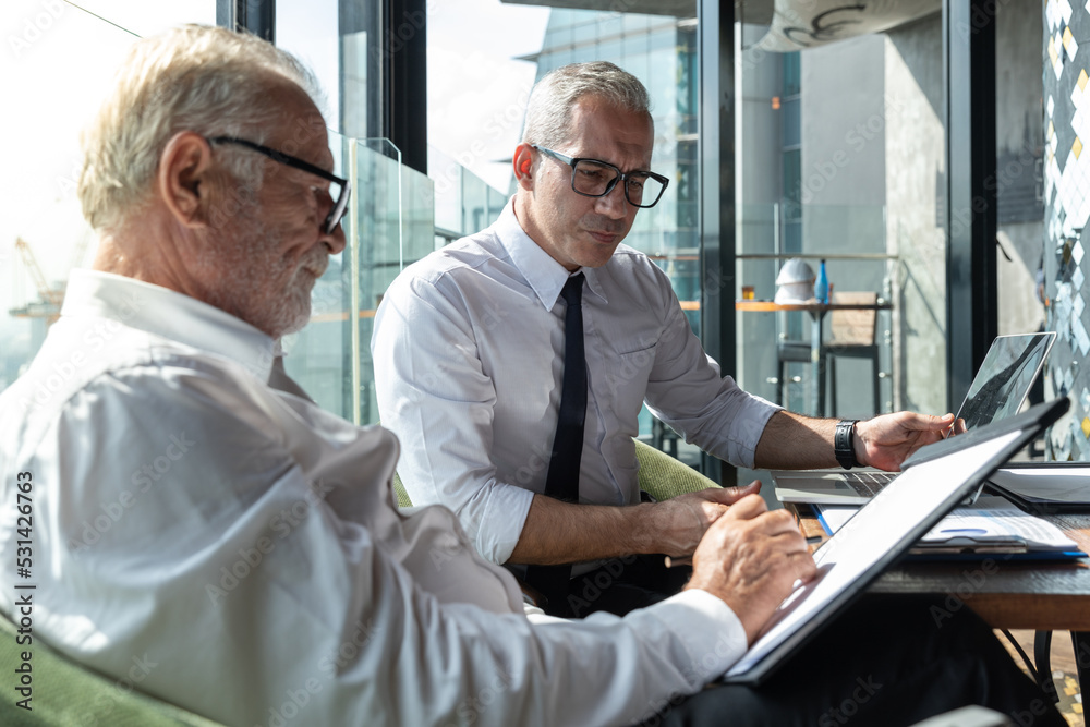 Two business men discussing idea in the office. One man is explaining while the other is listening. Both are wearing white shirt. The younger man is wearing tie and glasses.