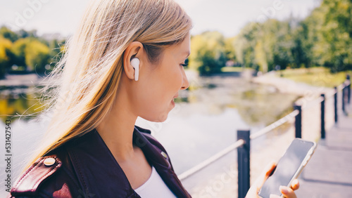 Young caucasian girl with wireless headphones in the park using tablet, phone and smiling