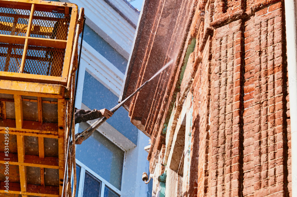 Obraz premium Water jet cleaning of facades. High pressure washing of the facade of a historic building using a hydraulic lift. A worker directs a jet of water at a red brick surface. Splash cloud. Bottom view.