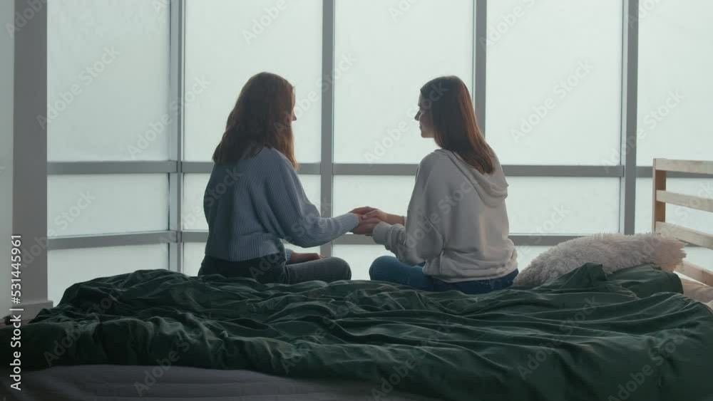 Girlfriends, Students or Sisters Back View Sitting on Bed Near Panoramic Window Supporting Each Other Holding Hands Warm Relation. Adolescence Concept. Mental Health.Friendly Support. Girlfriends, Students or Sisters Back View Sitting on Bed Near Panoramic Window Supporting Each Other Holding Hands Warm Relation. Adolescence Concept. Mental Health.Friendly Support.