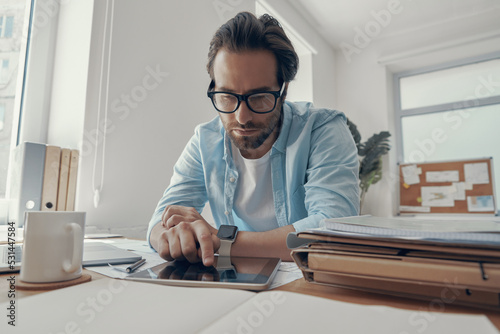 Handsome young man working on digital tablet while sitting at his working place in office © gstockstudio