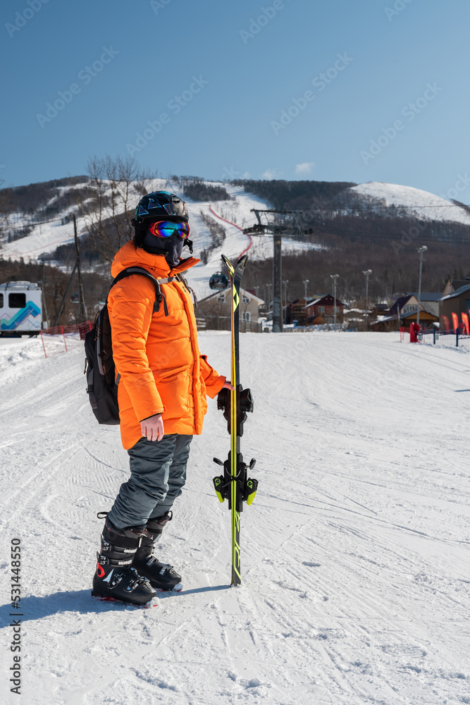 A skier in an orange jacket stands in front of a ski slope in full gear and skis in his hands