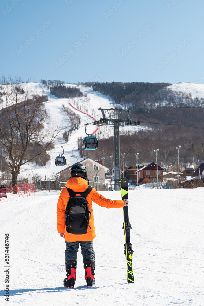 A skier in an orange jacket stands in front of a ski slope in full gear and skis in his hands