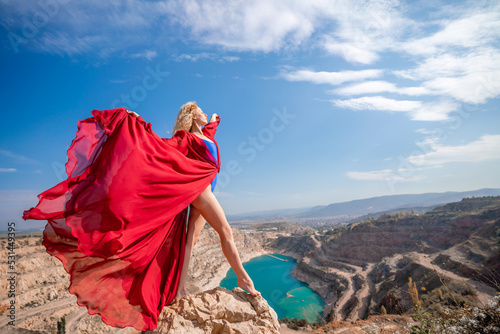Side view of a beautiful sensual woman in a red long dress posing on a rock high above the lake in the afternoon. Against the background of the blue sky and the lake in the form of a heart