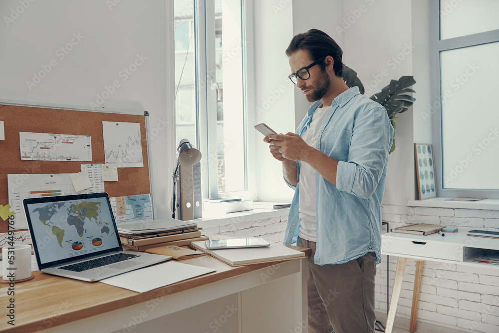 Confident young man using smart phone while standing near his working place in office