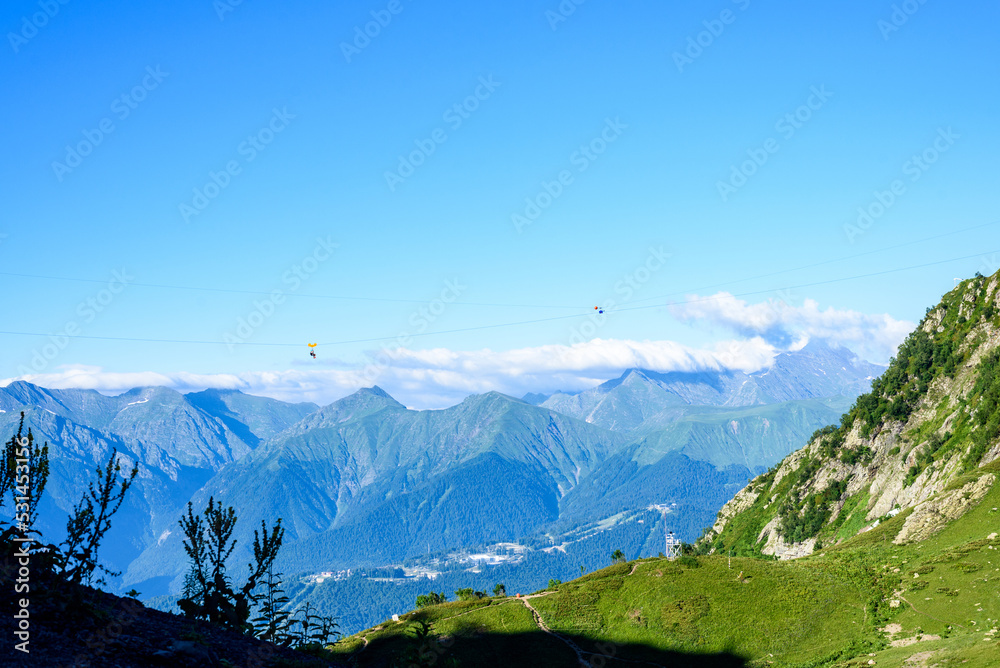 Misty mountains covered with white clouds against blue sky. Green hill slope with blossoming plants illuminated by bright sunlight