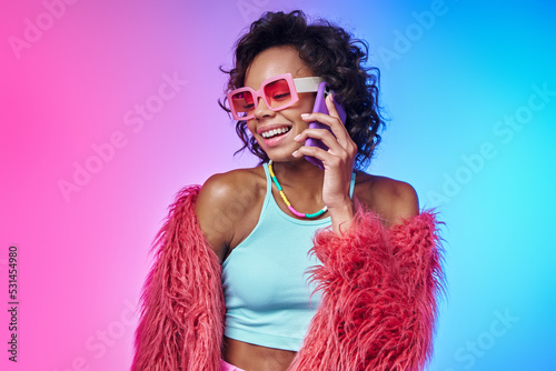 Happy African woman in trendy clothes talking on mobile phone against colorful background © gstockstudio