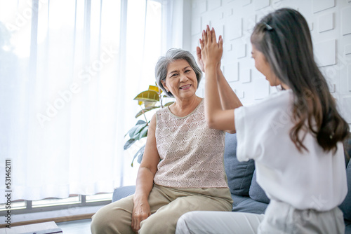 Asian grandmother and granddaughter talking and hi-five at home. Senior and young woman smiling and happy mood on sofa in living room.