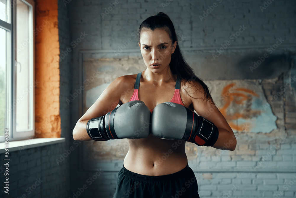 Naklejka premium Confident young woman in boxing gloves looking at camera while standing in gym