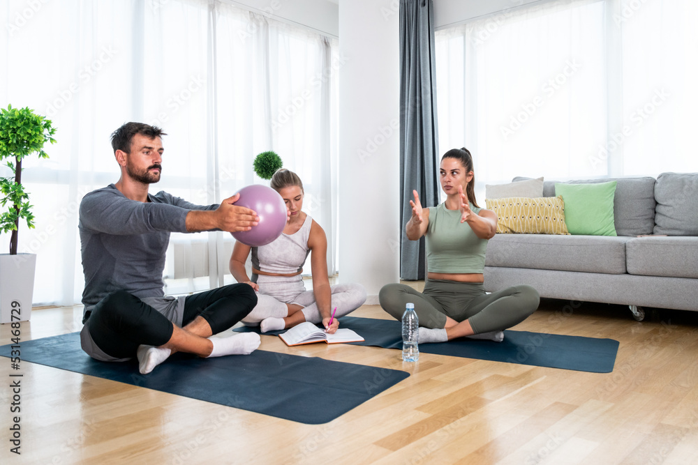 Young woman fitness and yoga instructor helping couple in their home for workout exercises. Female coach working with clients in their apartment. Husband and wife paying physical therapist for sport. Young woman fitness and yoga instructor helping couple in their home for workout exercises. Female coach working with clients in their apartment. Husband and wife paying physical therapist for sport.