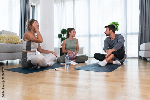 Young woman fitness and yoga instructor helping couple in their home for workout exercises. Female coach working with clients in their apartment. Husband and wife paying physical therapist for sport.