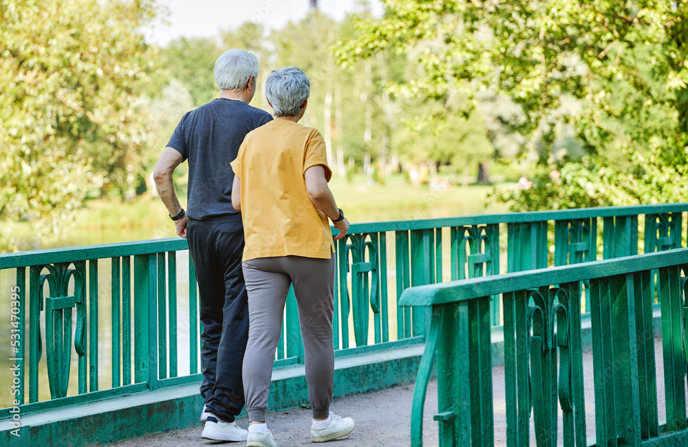 Rearview middle-aged wife and husband wear activewear strolling along bridge