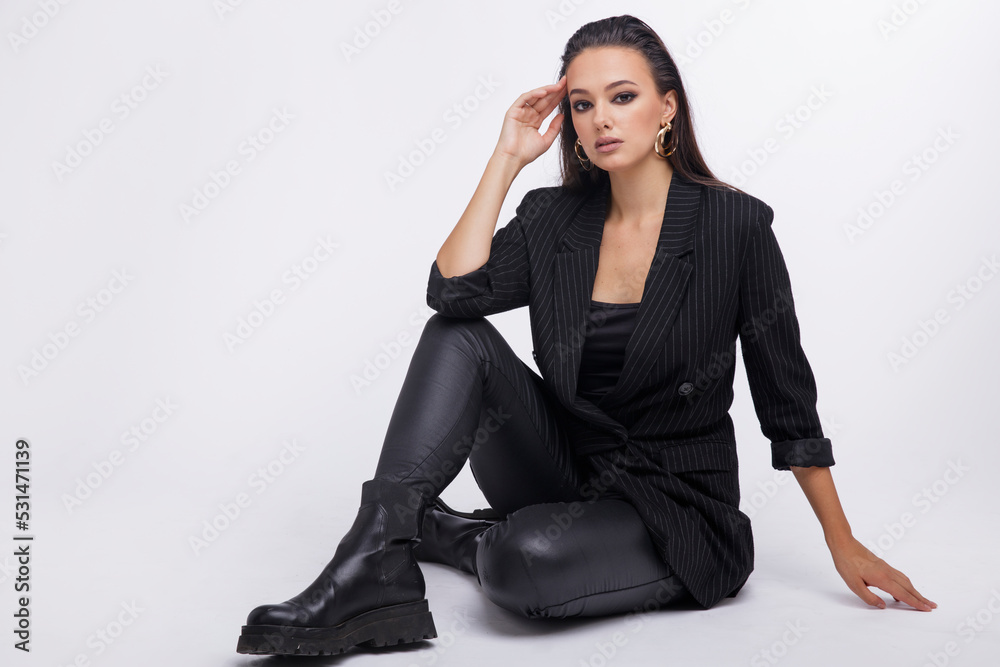Fashion photo of a beautiful elegant young woman in a pretty black jacket, leather pants, boots, posing over white background. The hair is gathered back, dark brunette. Studio Shot