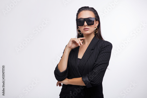 Fashion photo of a beautiful elegant young woman in a pretty black jacket, leather pants, boots, sunglasses posing over white background. The hair is gathered back, dark brunette. Studio Shot