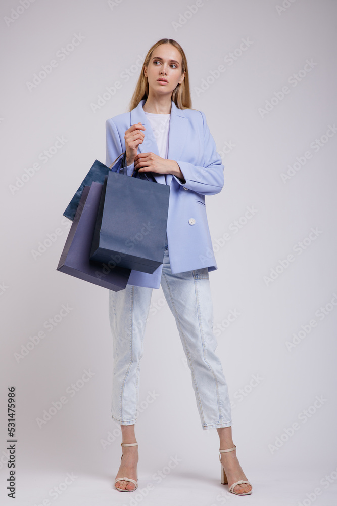 Fototapeta premium High fashion photo of a beautiful elegant young woman in a pretty blue jacket, blazer, denim jeans, shopping bags posing over white, soft gray background. Studio Shot. Slim Figure, Blonde