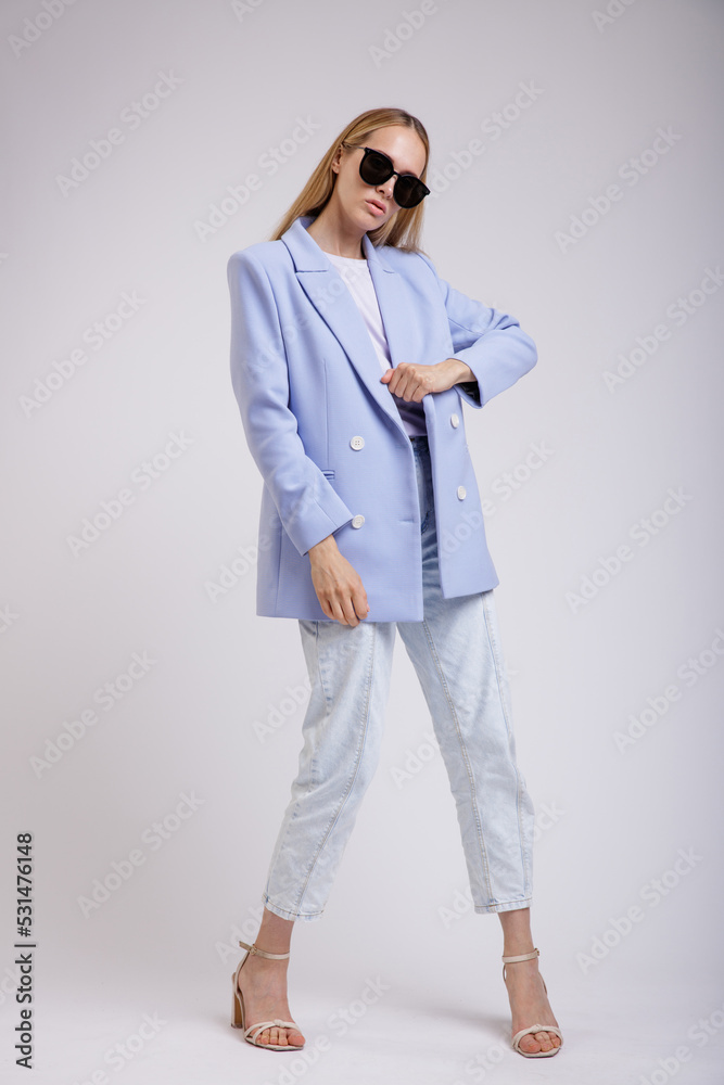 High fashion photo of a beautiful elegant young woman in a pretty blue jacket, blazer, denim jeans, stylish sunglasses posing over white, soft gray background. Studio Shot. Slim Figure, Blonde High fashion photo of a beautiful elegant young woman in a pretty blue jacket, blazer, denim jeans, stylish sunglasses posing over white, soft gray background. Studio Shot. Slim Figure, Blonde