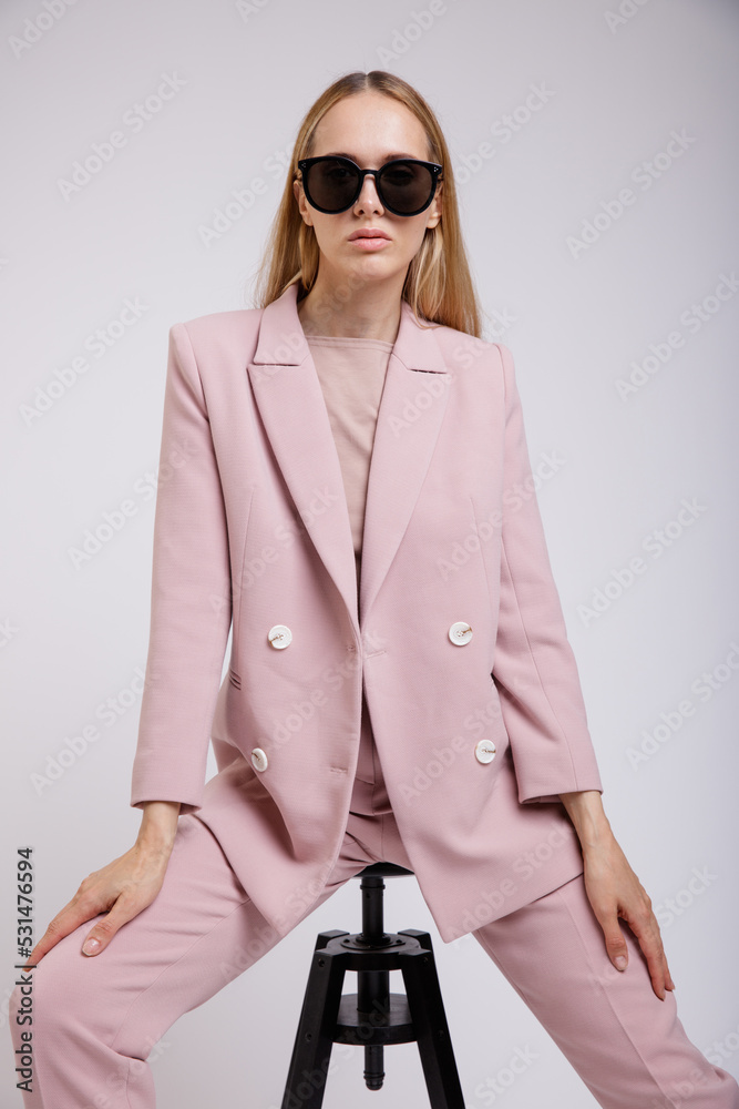 High fashion photo of a beautiful elegant young woman in pretty pink suit, jacket, blazer, trousers, pants, stylish sunglasses posing on white, soft gray background. Model sits on black chair High fashion photo of a beautiful elegant young woman in pretty pink suit, jacket, blazer, trousers, pants, stylish sunglasses posing on white, soft gray background. Model sits on black chair