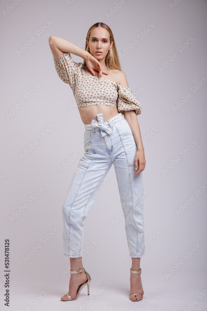 High fashion photo of a beautiful elegant young woman in pretty blue denim jeans, beige top with floral pattern posing on white, soft gray background. Slim Figure, Blonde. High fashion photo of a beautiful elegant young woman in pretty blue denim jeans, beige top with floral pattern posing on white, soft gray background. Slim Figure, Blonde.