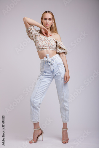 High fashion photo of a beautiful elegant young woman in pretty blue denim jeans, beige top with floral pattern posing on white, soft gray background. Slim Figure, Blonde. 