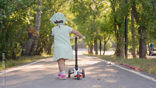 A beautiful 2-year-old girl pushes a scooter along an alley in the park, back view