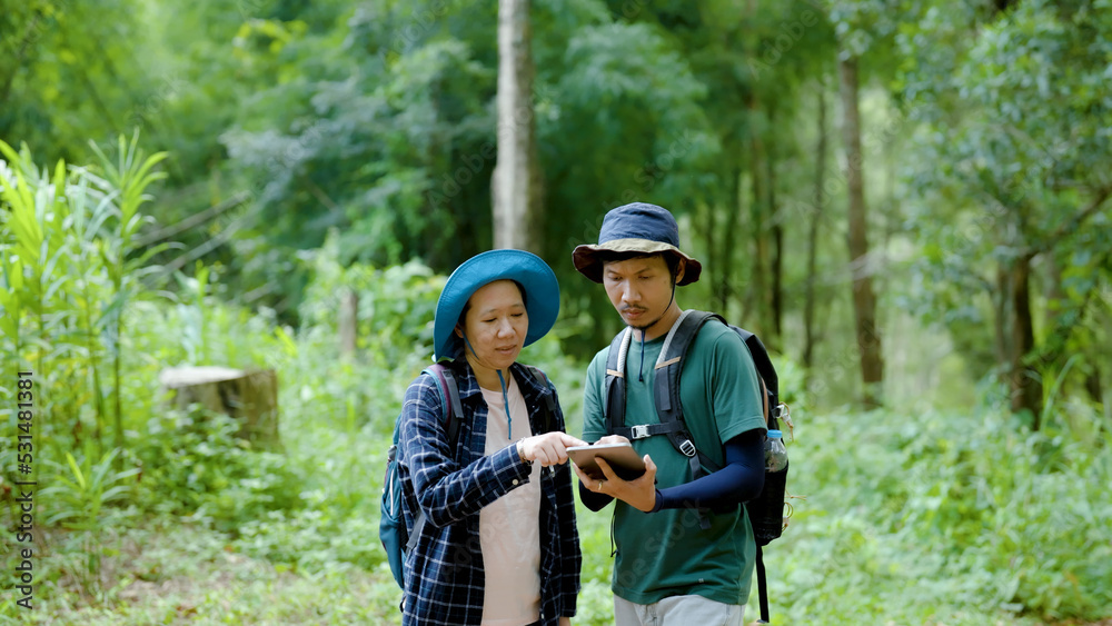 Young Asian tourist looking at a trekking map from a tablet in hand. and his lover to help him see the direction of walking up the mountain together. to see the path to travel further in the forest Young Asian tourist looking at a trekking map from a tablet in hand. and his lover to help him see the direction of walking up the mountain together. to see the path to travel further in the forest