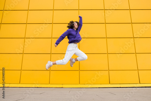 Happy positive woman in purple hoody have fun on yellow mall wall. Millennials girl with good mood flying in a jump isolated on colorful background