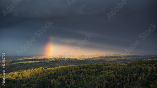 The sun shines through a gap in the cloudy sky and brings the end of the rainbow to light