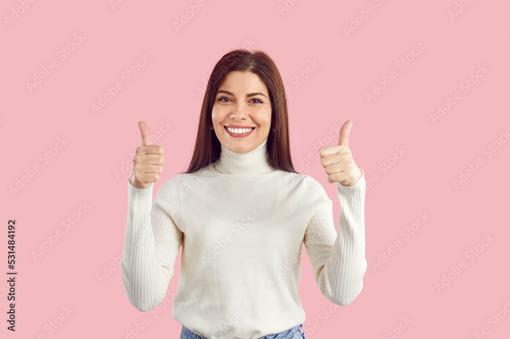 Portrait of happy beautiful young brunette Caucasian woman wearing casual white turtleneck standing isolated on pastel pink background, looking at camera, showing thumbs up sign and smiling