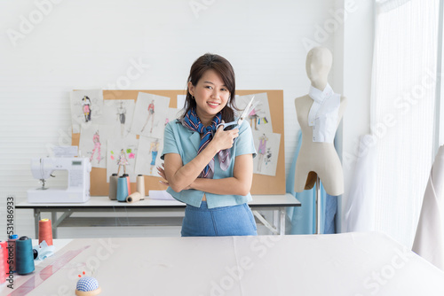 Portrait of happy asian female dressmaker holding scissors in the sewing workshop. Smiling woman seamstress working in tailor shop. Fashion  dressmaking or tailoring concept