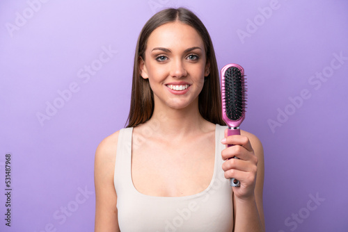 Young caucasian woman holding hairbrush isolated on purple background smiling a lot