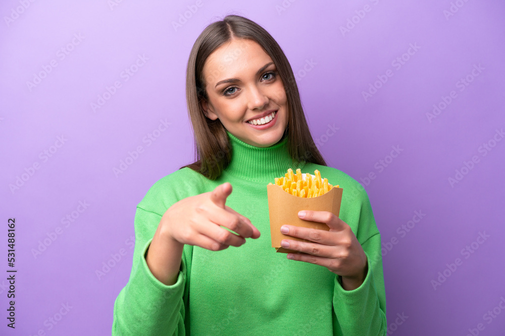 Young caucasian woman holding fried chips on purple background pointing front with happy expression Young caucasian woman holding fried chips on purple background pointing front with happy expression