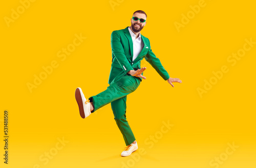 Cheerful guy in a stylish party outfit dancing in the studio. Full length portrait of a happy man wearing a fashionable green suit and sunglasses dancing isolated on a bright yellow color background photo