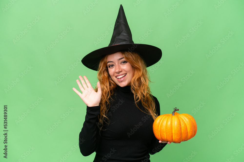 Young caucasian woman costume as witch holding a pumpkin isolated on green screen chroma key background saluting with hand with happy expression Young caucasian woman costume as witch holding a pumpkin isolated on green screen chroma key background saluting with hand with happy expression