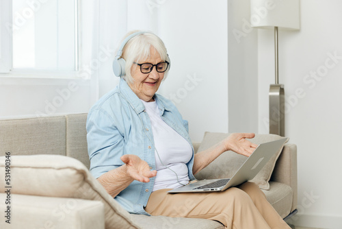 a joyful elderly woman is sitting on a cozy sofa talking via video link keeping in touch with loved ones at a distance talking to them through headphones © Tatiana