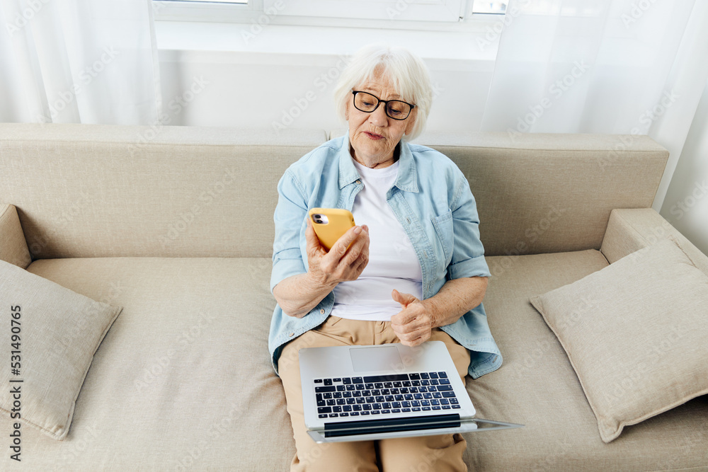 an annoyed woman is sitting at home on a comfortable sofa with a laptop on her lap and looking at her smartphone while gesturing with her hand. The concept of working from home